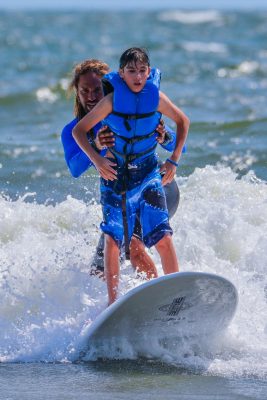 Athlete and coach on a surf board riding a way. The coach is on the back of the board holding the waist of the athlete in front. The athlete is wearing a blue life jacket. The tip of the surfboard is pointed up out of the water. 