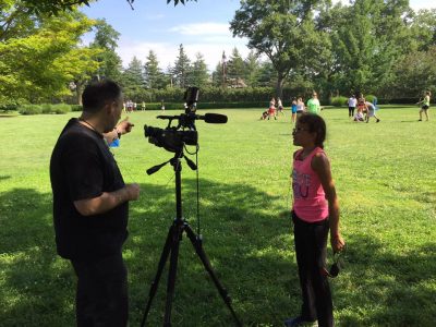 Photo taken in a large green park area. Camera man is pointing with a large video camera in front of him. He is interviewing an athlete who is standing in front of the camera speaking to it. Lots of athletes and coaches playing in the background. 