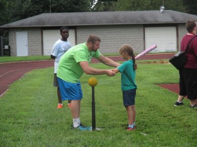Athlete holding a bat facing a beep ball balancing on a batting tee. Her coach is standing in front of her, using his hand over hand guidance to help her position the bat.