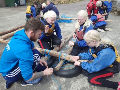 Four athletes and coaches working together to build a float. One athlete is tying blue rope around a log that is balancing on a tire. Another group of athletes and coaches are working on two other corners of the float in the background. 