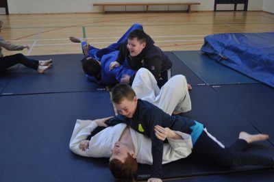 Two pairs of athletes and coaches on large blue mats. Athletes are pinning their coaches down on the mat using a judo hold. Athletes are smiling big. 