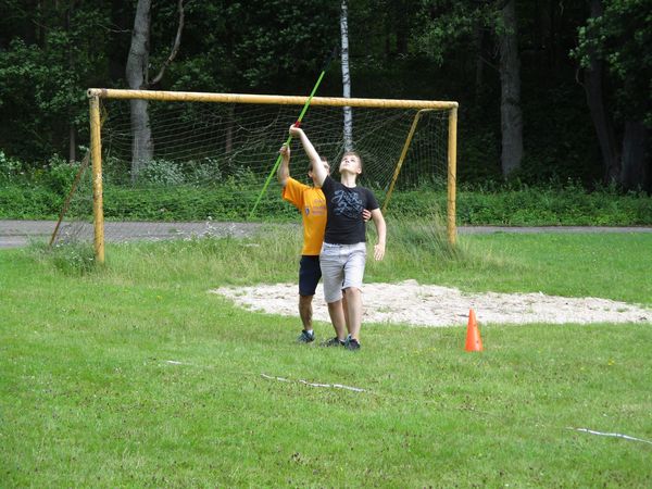 Coach providing hand over hand guidance to athlete holding javelin. Both have one hand on the javelin which is pointed in the air above their heads. Yellow soccer net in the background. Both are looking up to the sky.