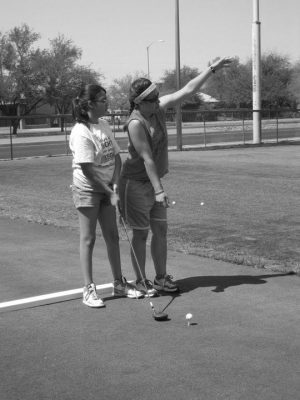 Maria working with an athlete in golf. Maria holding her arms open to show the distance of the ball to the athlete who is standing next to her holding a golf driver. Photo is greyscale.