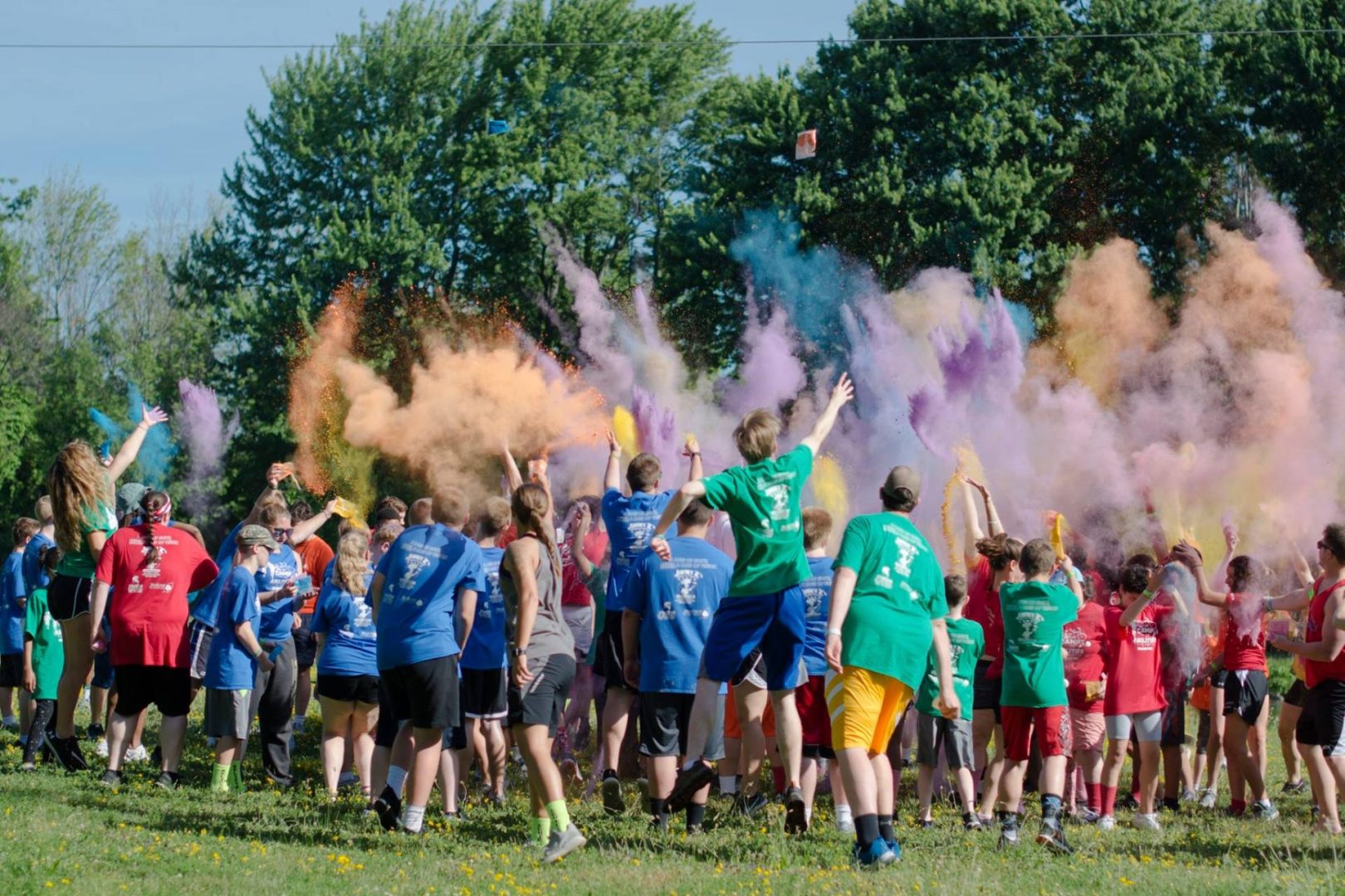 Camp Abilities Brockport Athletes and Coaches celebrating with color bust. Large group of athletes wearing blue, green and red camp tshirts throwing orange, purple and blue dust in the air.