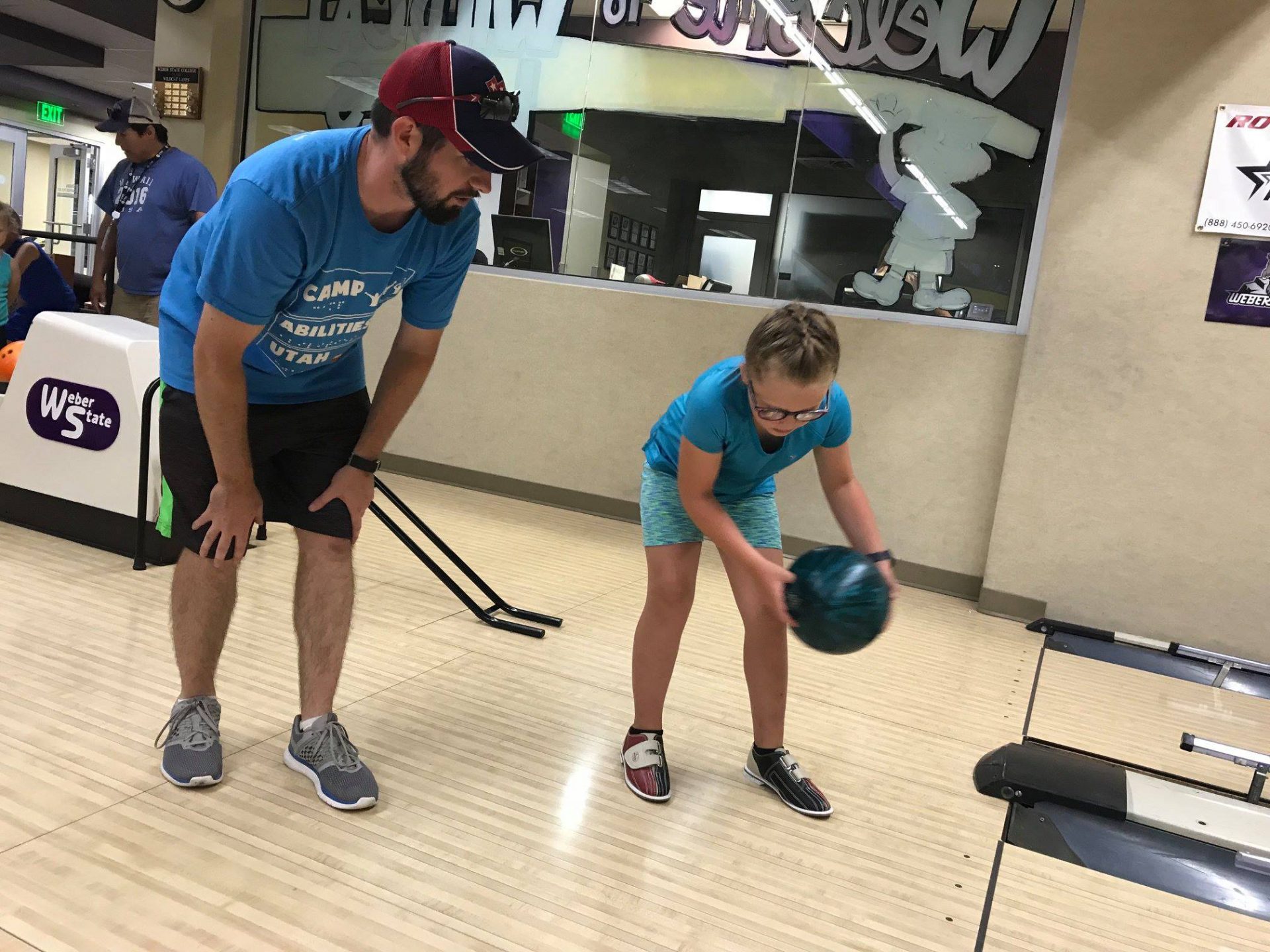 Photo taken from the side. An athlete is holding a large blue bowling ball with two hands. She is positioned in front of the bowling lane. Her coach is standing behind her with his hands on his knees awaiting her throw.