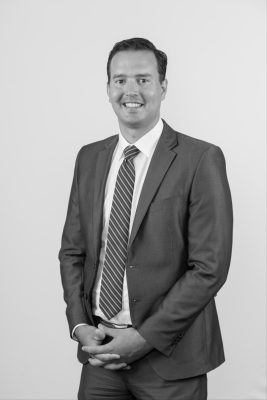 Photo of Corey smiling for a headshot photo. His arms are folded at his waist and he is wearing a suit and tie.