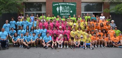 Group photo of Camp Abilities Brockport. All children and coaches lined up by Color Team (Blue, Pink, Yellow, Orange) with Sport Specialists and Staff in bright green. All people smiling at the camera.
