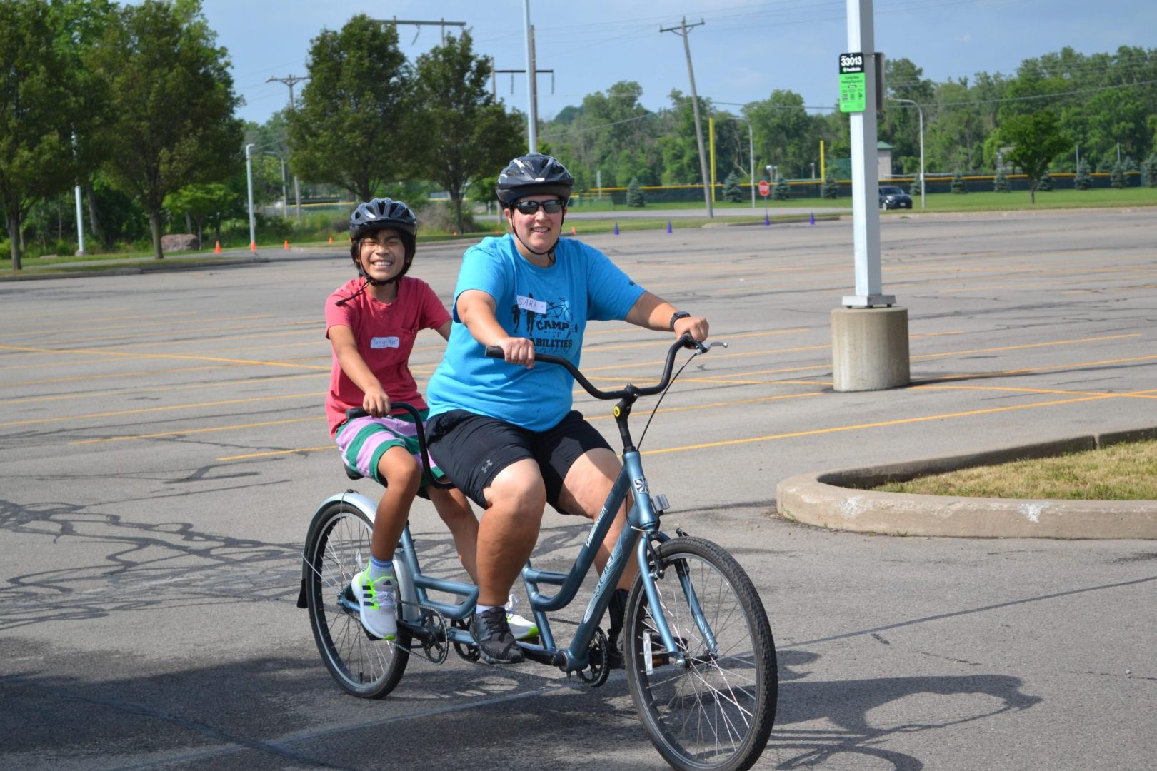 Coach and athlete riding a tandem bike. Coach is smiling wearing a blue shirt with black shorts and athlete is smiling wearing a red shirt with striped pink and blue shorts.