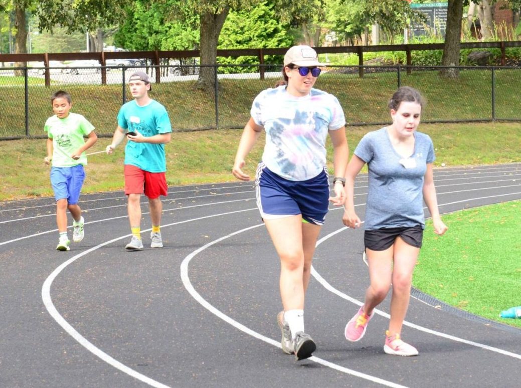 Athletes and coaches running around the track holding a tether. Coach in foreground is wearing blue tie-dye shirt with sunglasses, a hat, and blue shorts guiding an athlete wearing a blue shirt, black shorts, and has brown hair. Coach in the background is wearing a bright blue shirt with red shorts and athlete is wearing a green shirt with blue shorts.