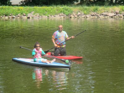Athlete sitting on blue paddle board in the water. Coach kneeling on red paddle board next to the athlete. Both individuals are smiling at the camera while holding their paddles. Rock shore and grass in the background. 