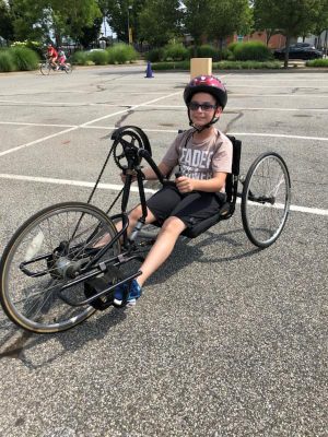 An athlete sitting in a handcycle bike that is low to the ground. He is parked in a parking lot. Another athlete and coach are riding a tandem bike in the background. The boy is smiling for the camera wearing his bike helmet. 