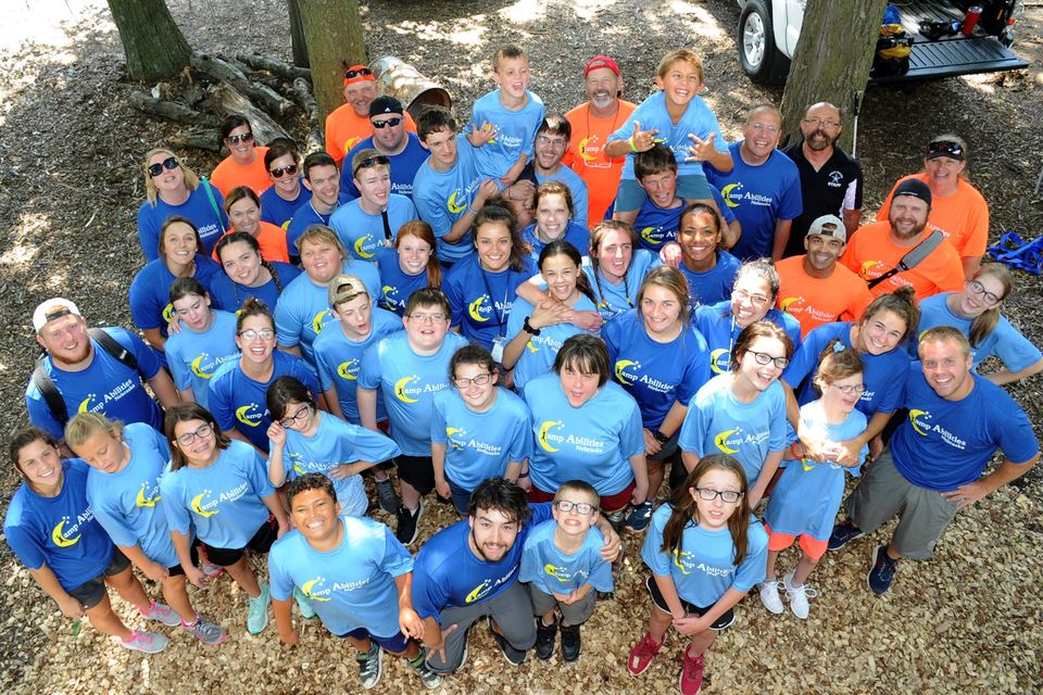 Group photo of athletes and coaches standing in the woods looking upwards, smiling at the camera. Everyone is wearing a blue or orange camp tshirt.
