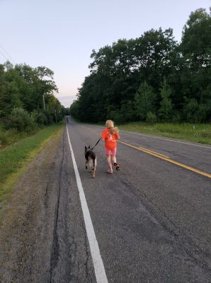 Athlete running on paved road with a dog. Athlete and dog have their backs to the camera. Athlete is holding the leash. Lots of green trees on both sides of the road. The road goes on into the distance as far as the photo can see. 