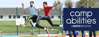 An athlete and coach jumping the long jump in mid air. Both have one leg stretched out in front and the other leg behind them. Both have their arms swinging up in the air. 3 spectators standing on the side. Camp Abilities - A Cara Initiative is written in white letters covering the 3 spectators. 