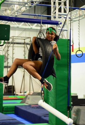 Athlete swinging on a tire swing in mid air. Athlete is in the middle of an obstacle course with lots of obstacles hanging in the background. She is reaching to grab the next obstacle while she swings from the tire. 
