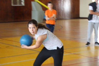 Athlete is in a gymnasium holding a blue goalball in mid swing. She is looking in front of her with the goalball pulled back behind her. She is in the motion of the throwing the goalball with two hands. Two other athletes standing in the background with their arms crossed. 