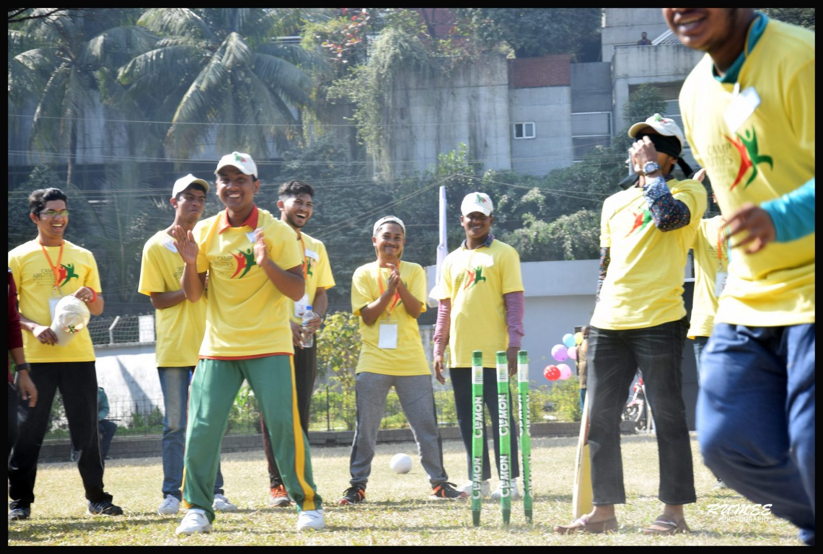 Group of Bangladesh athletes and coaches smiling, clapping, and cheering while playing cricket. Trees and houses in background. Standing in a field with green cricket wickets in ground.