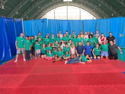 Camp Abilities Brazil Group Photo. Large group of athletes and coaches. Most are wearing green camp tshirts. Two rows are kneeling on a large red mat. Back rows are standing on red mat. Blue curtains hanging in the background with large dome window. 