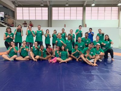 Camp Abilities Brazil group photo of 28 athletes and coaches. Front row is sitting on a large blue mat and back row is standing. Athletes and coaches are holding their two fists up in front of their faces. Wearing green camp tshirts. 