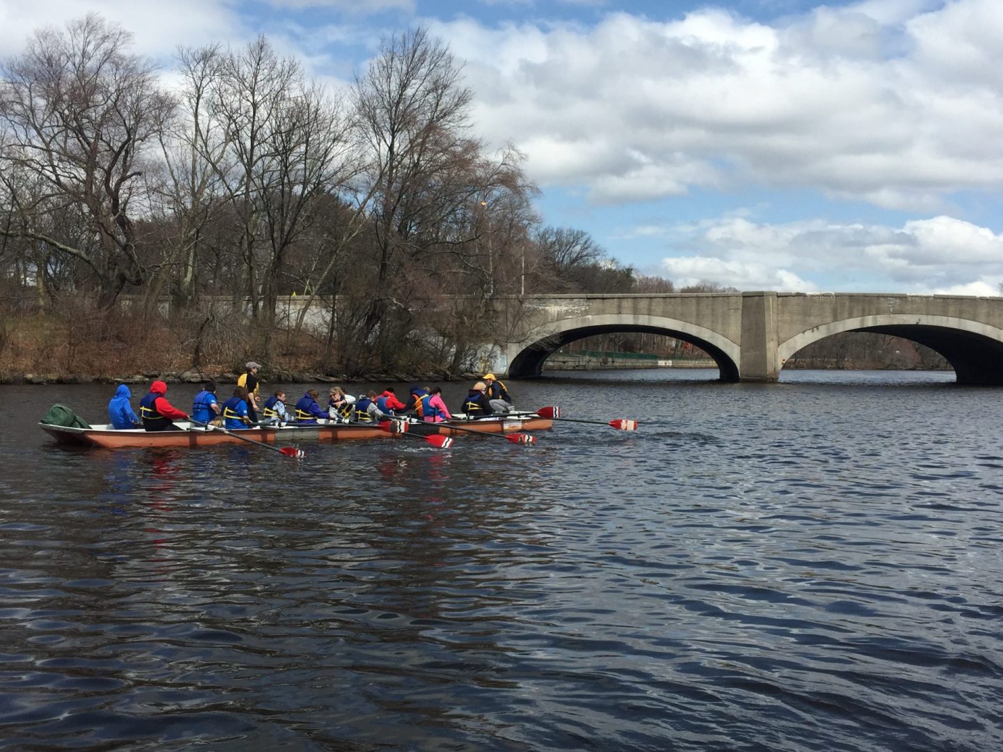 Large red canoe in water with several athletes and coaches on board. Paddling towards bridge with two large passages to move through. Blue, cloudy sky with large, bare trees on the shore.