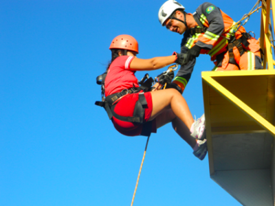 Athlete wearing helmet and harness on high ropes obstacle. Paused at the top of climbing wall. Climbing instructor wearing helmet and safety gear reaching down towards athlete.