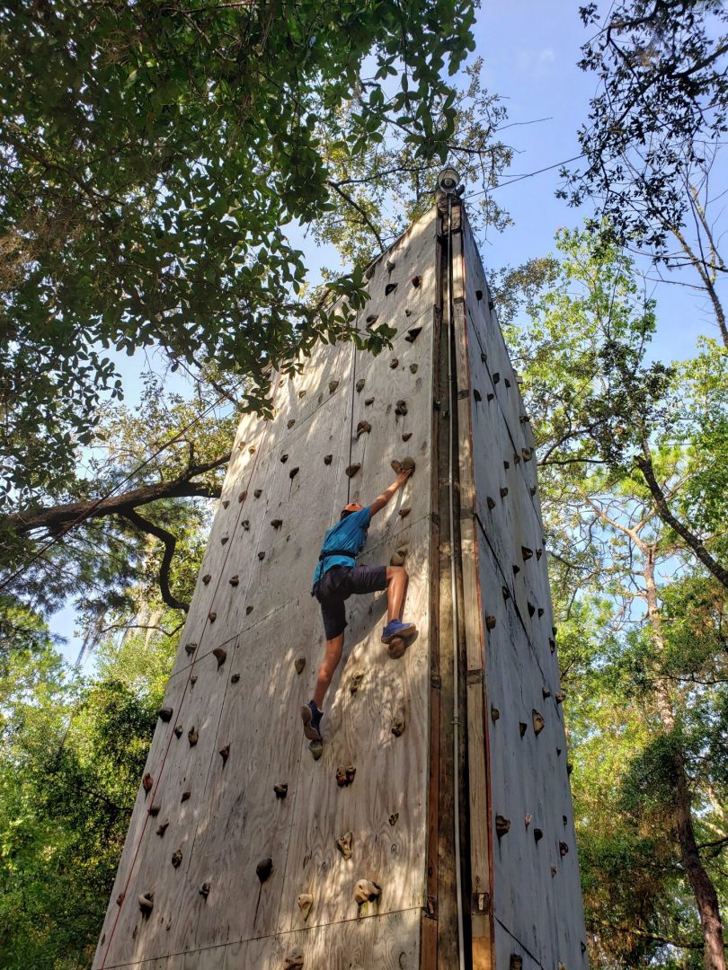 Athlete is half-way up the climbing wall. Wall is surrounded by woods. Athlete is wearing a helmet and harness. Their hands and feet are resting on the climbing wall handles.