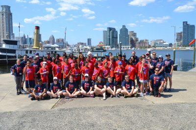 Group photo of athletes and coaches wearing red and navy blue tshirts. Everyone is smiling at the camera. Bright blue sky and many tall buildings in the background. Surrounded by water with a boat sitting in the background.