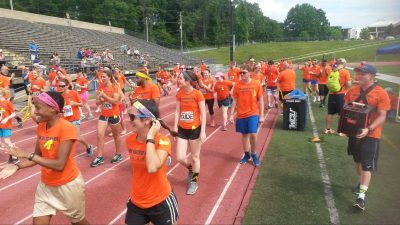Lots of athlete and coaches wearing orange tshirts. Smiling and walking on the track preparing to start their triathlon. Bleaches in the background with a few spectators.