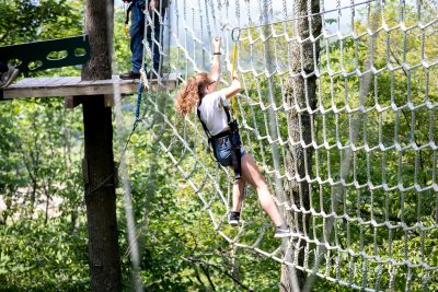 Photo taken in the woods with lots of greenery in the background. Athlete is wearing a harness and is positioned in the middle of a climbing net. The net is wrapped around trees to hold it in the air. The athlete has a wooden platform to her left where a coach is waiting for her to cross the net.