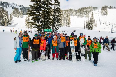 Large group photo with snowy trees and ski hills in the background. Athletes and coaches are wearing winter gear including helmets and orange vests that say guide and visually impaired. Some people are holding snowboards and skis. Some people are wearing ski/snowboard glasses.