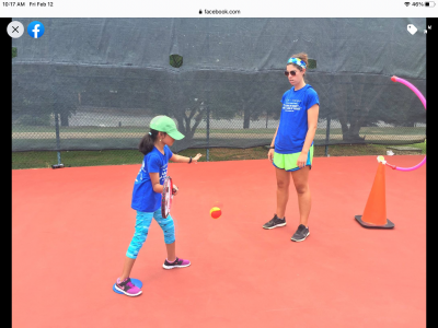 Athlete and coach standing on tennis court. Athlete is holding a tennis racket. She is mid swing, about to contact the ball that is in the air in front of her. There is a target in front of the athlete made of a pool noodle balancing on a large orange cone.
