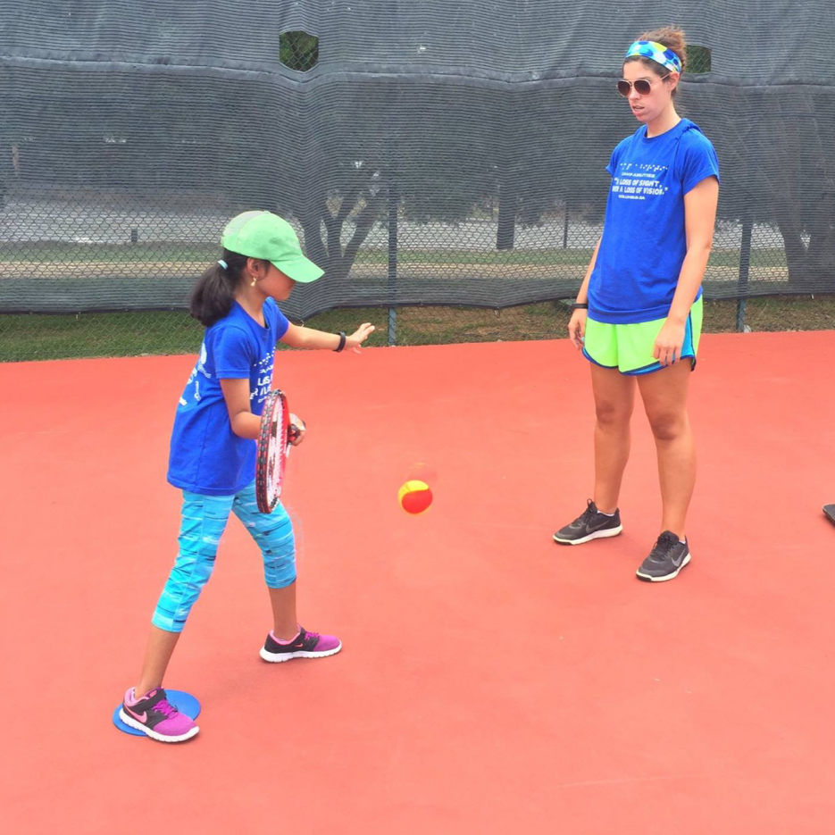 Athlete and coach standing on tennis court. Athlete is holding a tennis racket. She is mid swing, about to contact the ball that is in the air in front of her. There is a target in front of the athlete made of a pool noodle balancing on a large orange cone.