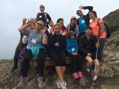 Group photo of athletes on a wooden platform in the middle of a large rock mountain. Athletes are making silly faces and holding their hands up by their heads. 