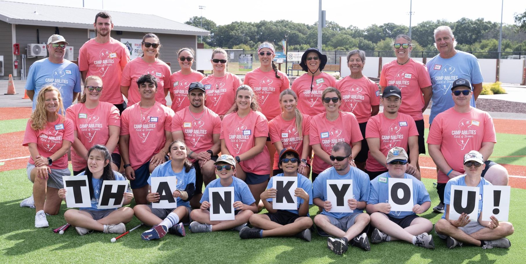 Group photo of Camp Abilities New Jersey - Field of Dreams. Coaches are in pink shirts and athletes are in blue shirts sitting in the front row of the picture holding individual letter signs spelling out "thank you".