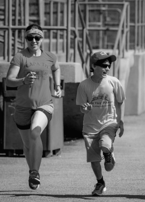 Maria running next to an athlete on the track smiling. Athlete is waering short sleeve shirt  and shorts looking off to the right. Maria wearing a short-sleeve shirt and shorts smiling running forward. Phot in greyscale.