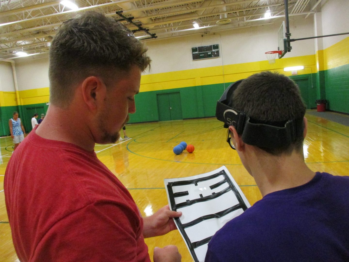 Athlete and Coach standing inside a gymnasium. Athlete is holding a tactile map while he and his coach go over the rules and dimensions of the goalball court.
