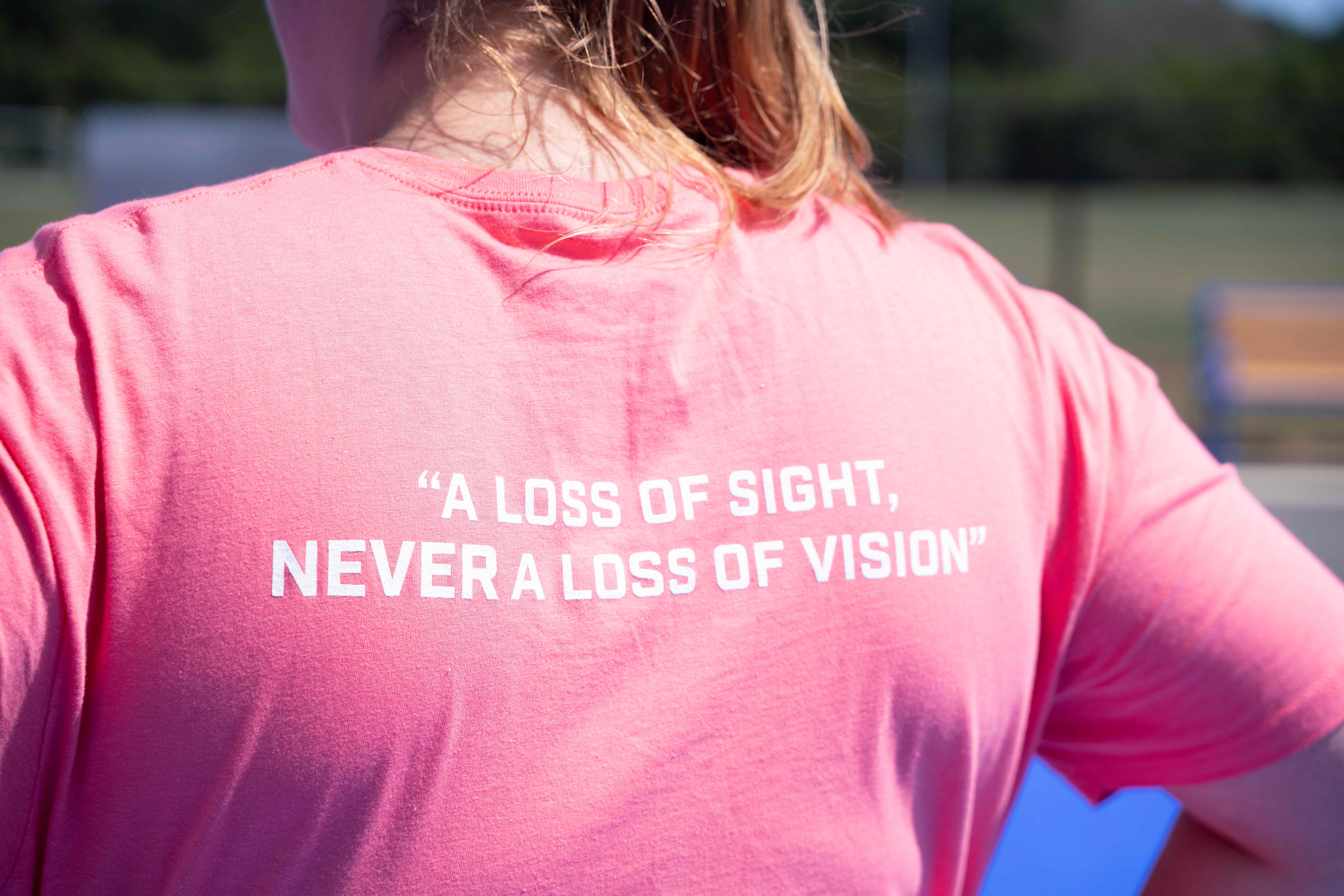 Photo of the back of a coach's shirt with the Camp Abilities motto: "A loss of sight, NEVER a loss of vision." Coach has blonde hair in a pony tail wearing a pink shirt with white writing.