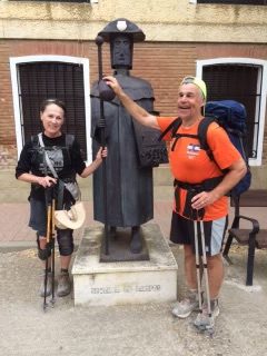 Terry Kelley and partner, Anne smiling while posing for photo with black metal statue. Both are wearing backpacks and holding walking poles. Brick building in the background. 