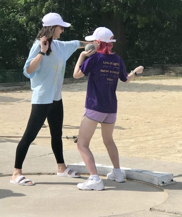 One coach wearing black leggings, blue long sleeve shirt, and white hat teaching an athlete how to throw a shot put. The athlete is wearing light purple shorts, purple shirt, and white hat and preparing to throw shot put forward in the ring.
