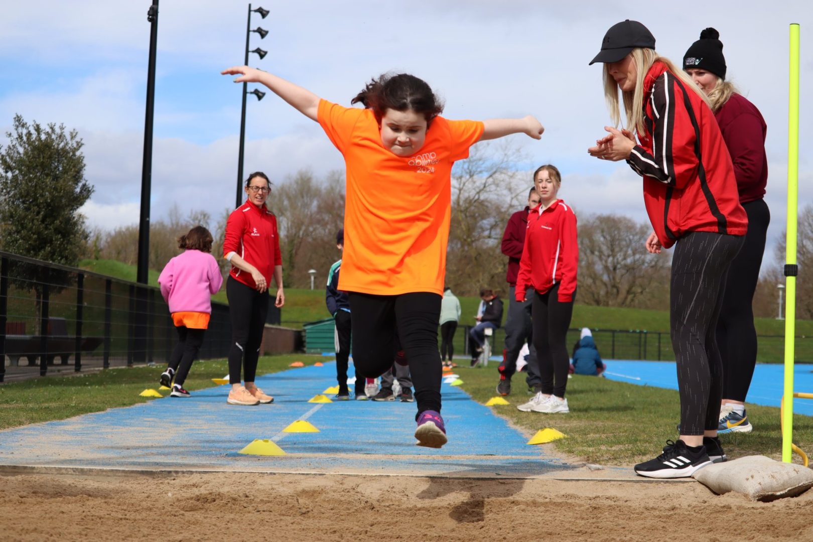 Photo of athlete in orange shirt and brown hair jumping into a sand pit for the long jump. Athlete is mid flight with arms stretched out to the sides like a T. Coaches standing on the side clapping to help athlete know when the jump.