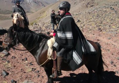 Terry Kelly sitting on a brown horse in full uniform including a helmet and riding boots. White horse and rider in the background. Brown, bare, mountain terrain.