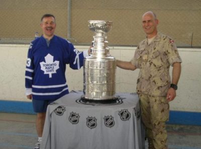 Terry Kelly smiling, posing next to the Stanley Cup with colleague. Both individuals have one hand touching the cup with rink boards in the background. 