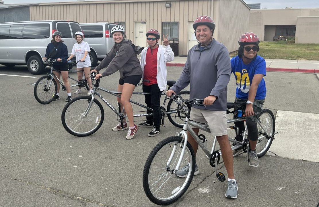 Three pairs of tandem bikes with coaches piloting and athletes on the back posing for photo prior to riding. All people are smiling, wearing helmets, and feet are on the ground.