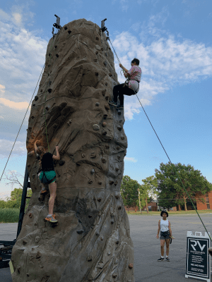 Two athletes climbing up opposite sides of a large rock climbing structure. The structure is temporality set up in a pavement area. Both athletes are harnessed in and are using their arms and legs to climb the wall. There is one spectator watching in the background. 