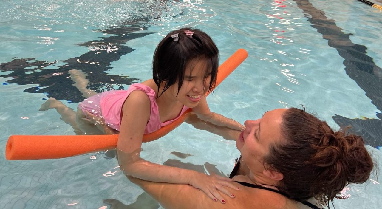 Athlete and coach swimming in the pool. Coach is holding the female athlete up in the water and athlete has a pool noodle supporting her underneath her waist to float. Athlete is wearing a pink bathing suit and is smiling while holding onto coaches shoulders.