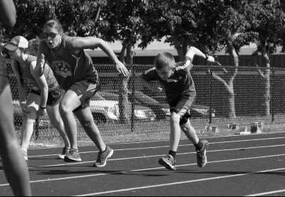 Alex running next to a male athlete on the track in greyscale. Photo taken as they began to run off the start line where both heads are ducked and arms are extended back to begin running.
