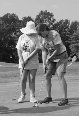 Alex teaching an athlete how to put in golf in greyscale. Alex is wearing short sleeves, shorts, and sunglasses looking at the ball. Athlete standing next to her is holding a putter and wearing a sun hat blocking their face.
