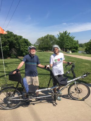 Paul and Susan smiling as they stand next to a tandem bike. Both are wearing helmets and are reading to ride the bike. Blue sky and green trees in the background. 