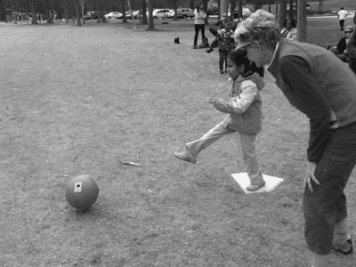 Judy teaching an athlete how to kick a ball for beep kickball. Judy is learning over with her hands on her knees as an athlete kicks the ball with her left leg at home plate. Photo is in greyscale.
