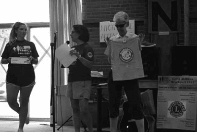 Judy standing with Lauren Lieberman and Kelsey from Camp Abilities Brockport Opening Ceremonies. Judy is holding a Beep Kickball shirt in her hands and Lauren is making an announcement on the microphone.